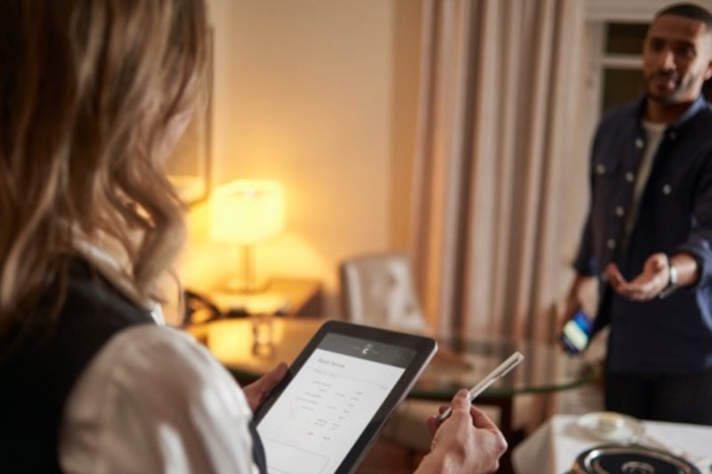 A person stands holding a tablet and pen and a man stands in front of them reaching out their hand