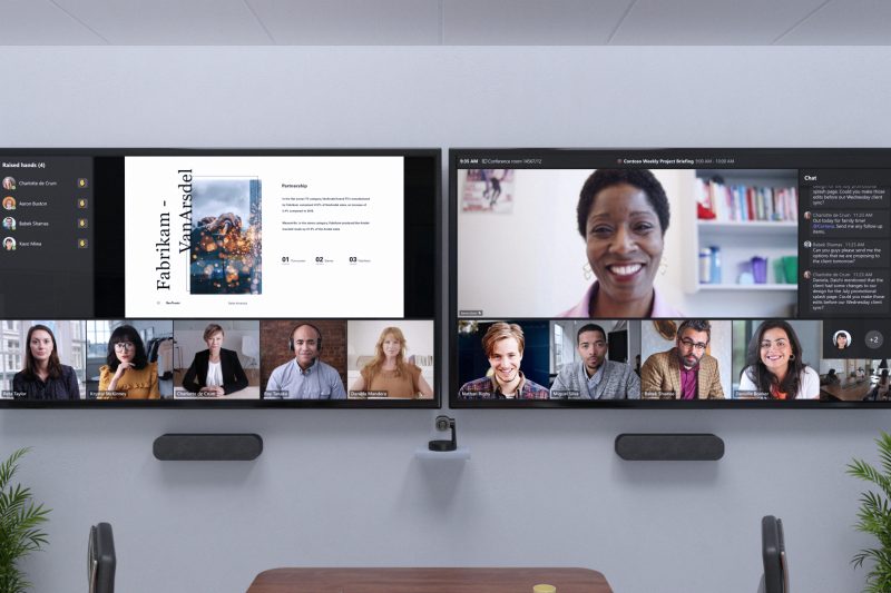 Remote attendees are across two front of room screens at eye level beside chat and a view of participants with raised hands.