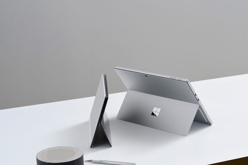 Two tablet computers with kickstands rest on a table next to a roll of duct tape and a precision cutting tool.