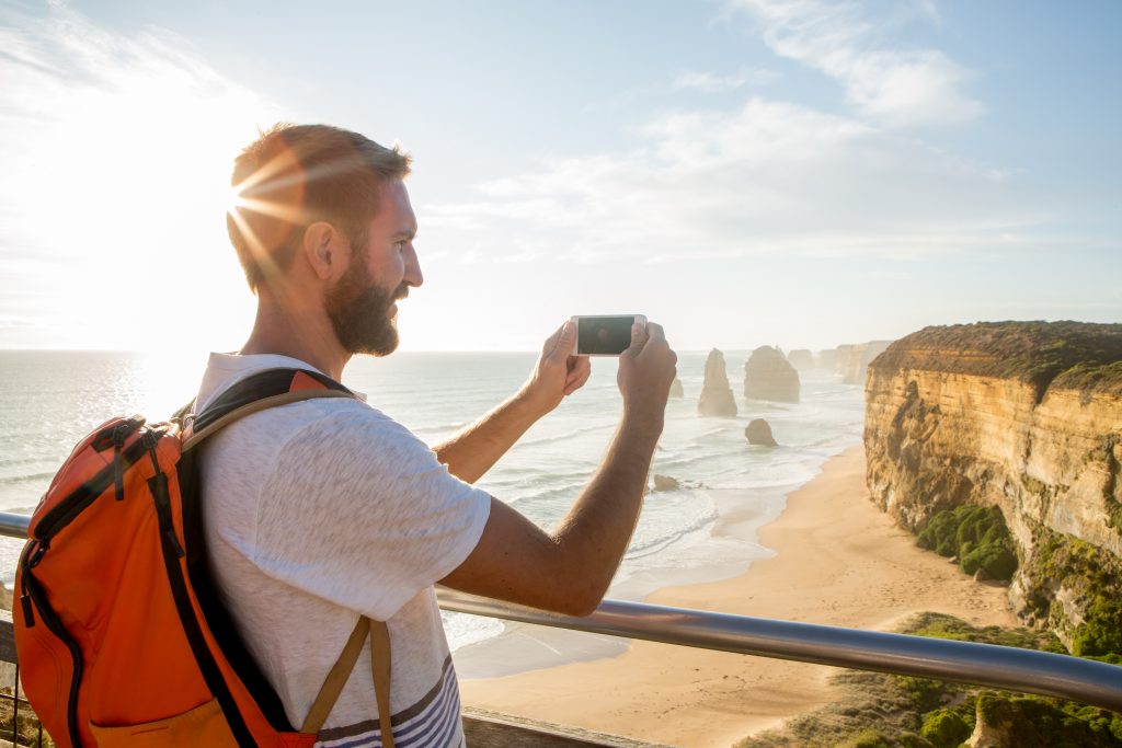 Cheerful young man takes a picture using a mobile phone of the Twelve Apostles sea rocks on the Great Ocean Road in Victoria's state of Australia.