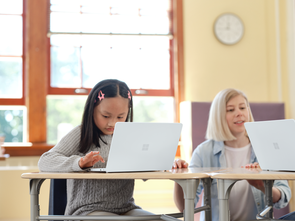 Elementary school age child sitting in a classrom with an open Surface Laptop SE in Glacier in front of her. A teacher is also shown.