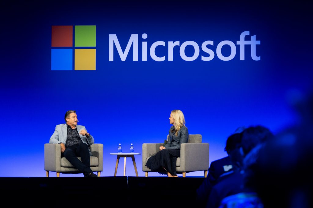A Caucasian male and female sitting across from one another in seated lounges, on stage, with a Microsoft logo behind them