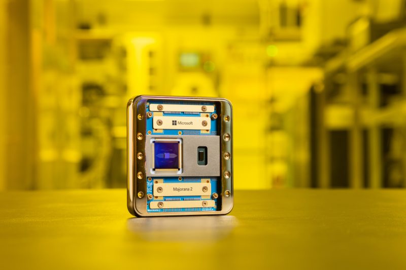 A mostly gold-colored chip with bits of blue showing stands on a table top against a yellow-hued lab background. The names "Microsoft" and "Majorana 2" are visible.