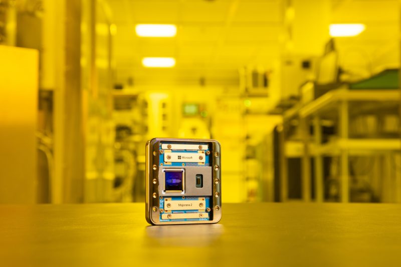 A mostly gold-colored chip with bits of blue showing stands on a table top against a yellow-hued lab background. The names "Microsoft" and "Majorana 2" are visible.