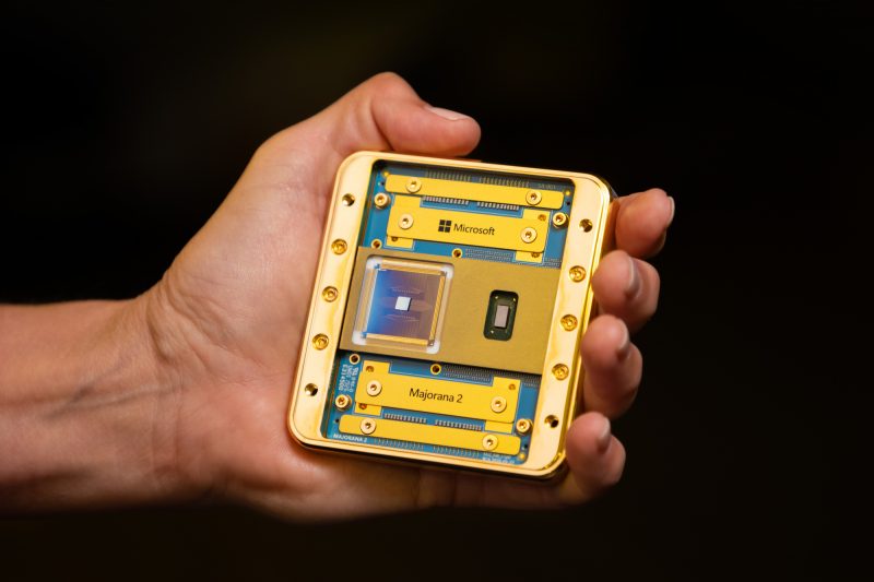 A mostly gold-colored chip with bits of blue showingis held in the palm of a person's hand. The names "Microsoft" and "Majorana 2" are visible.