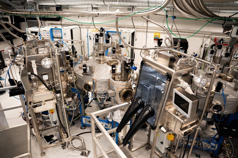 A large room with a tangle of wires and pipes along the ceiling, plus more wires and smaller pipes connecting various machines to the large, round quantum refrigerator in the center.