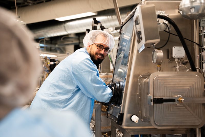 A bearded man with glasses, wearing a pale blue clean suit and a hair net, smiles at the camera as his arms are reaching into a sealed glass box for working with quantum materials.