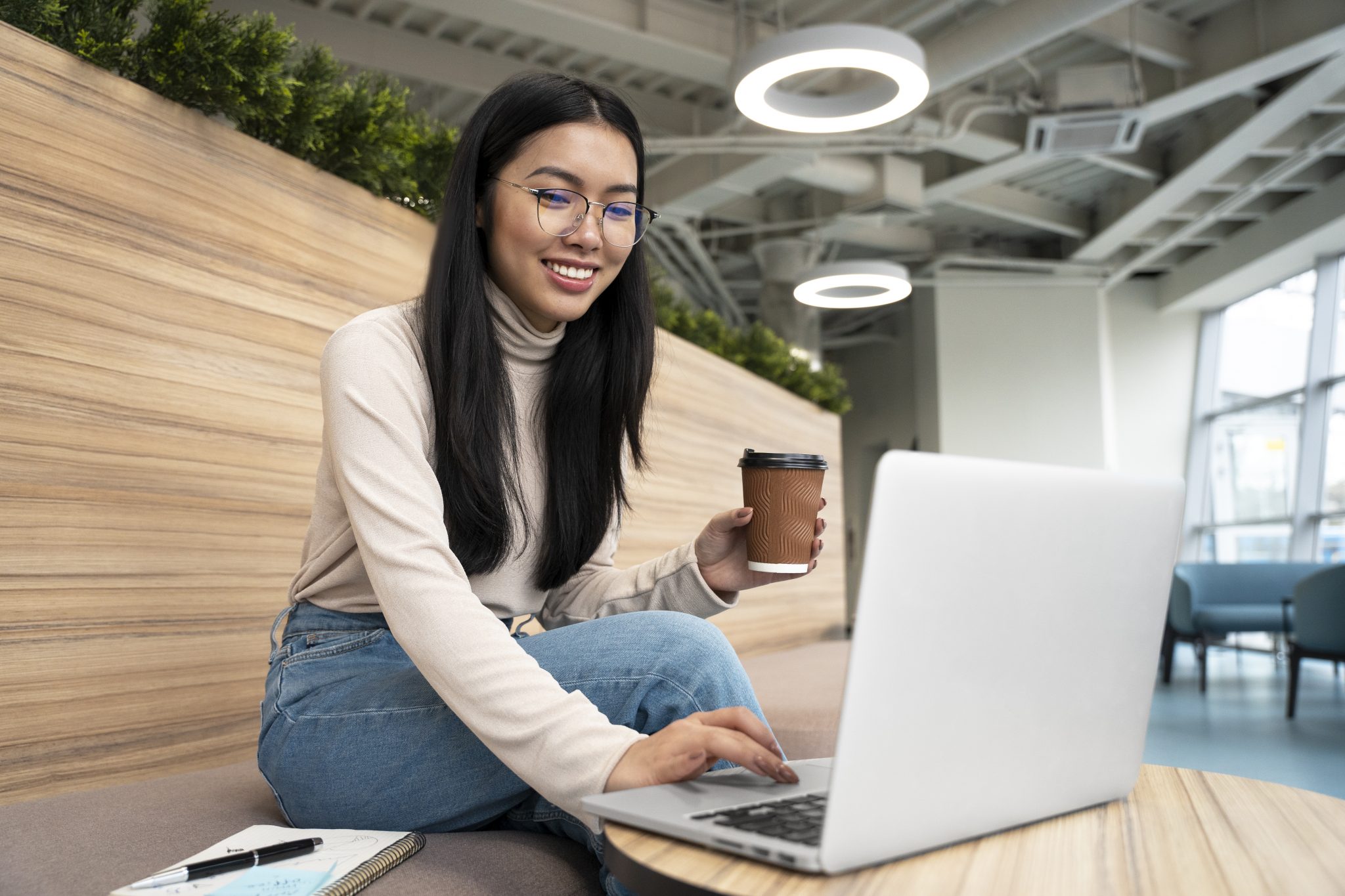 Asian girl with coffee cup in one hand and typing on a laptop with the other