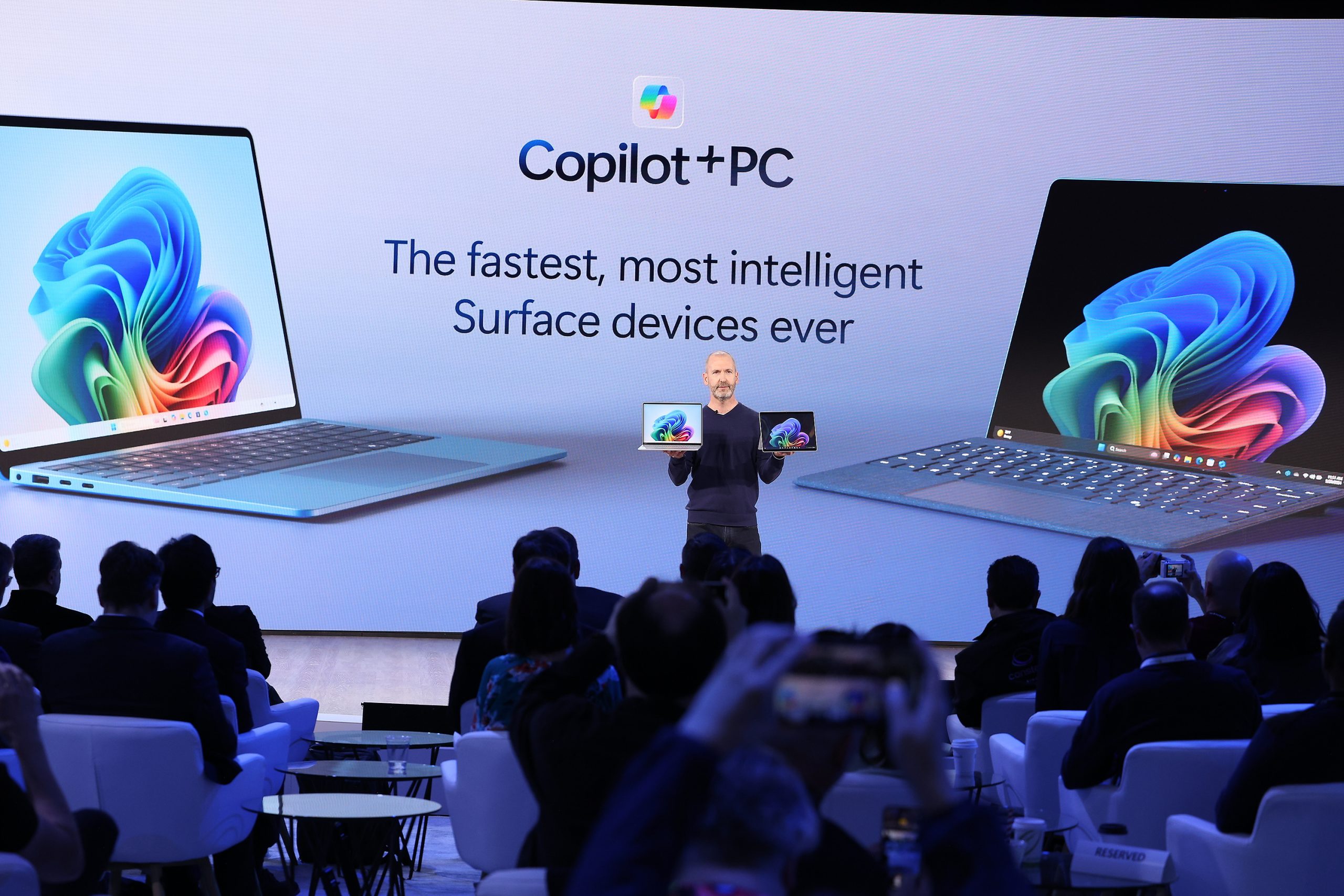A man standing on stage in front of an audience holding two Copilot+ PCs, in front of a screen that reads, "The fastest, most intelligent Surface devices ever
