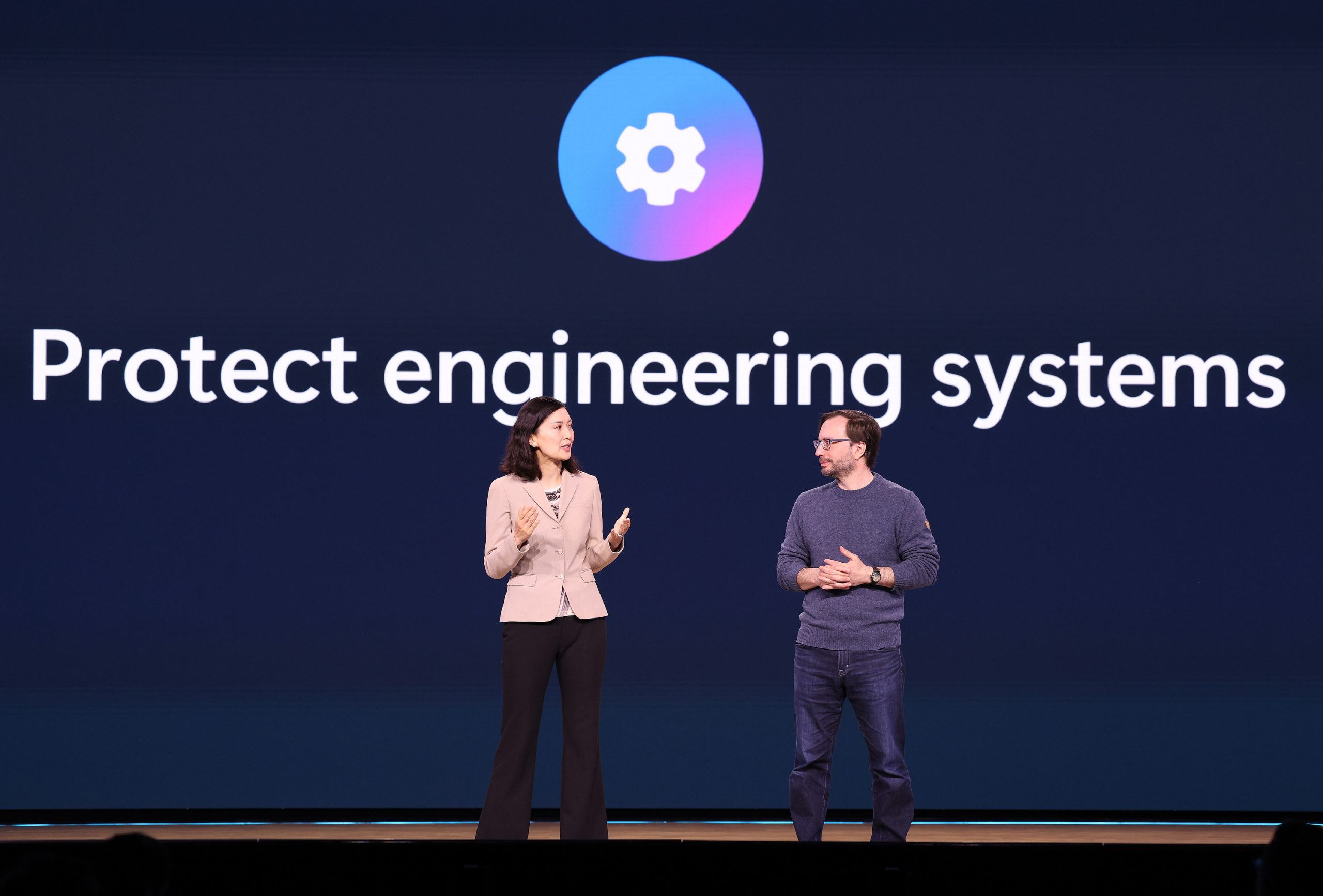 A man and woman standing on stage with text on a screen behind them that reads, "Protect engineering systems"