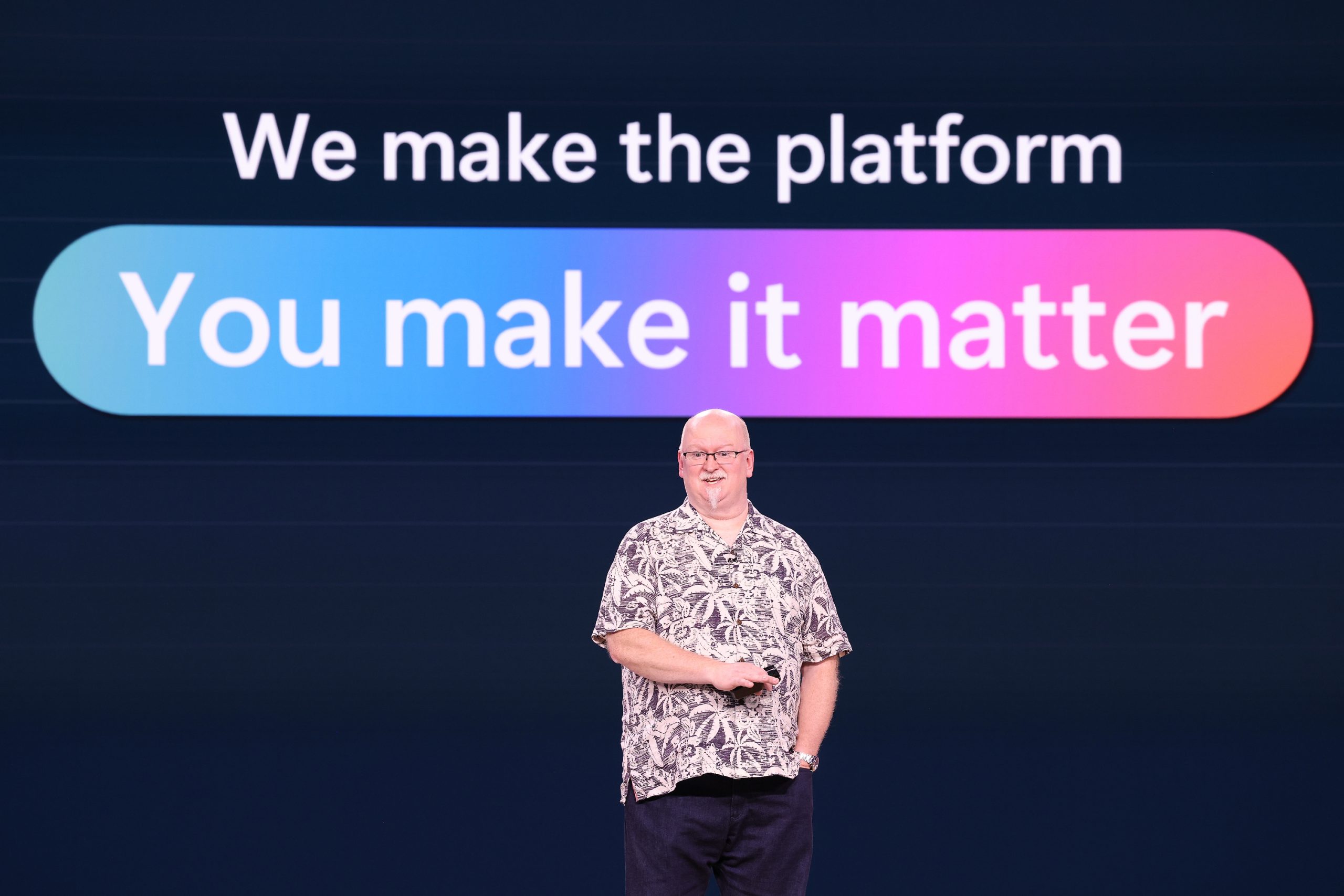 A man standing on stage with text on the screen behind him that reads, "We make the platform. You make it better."