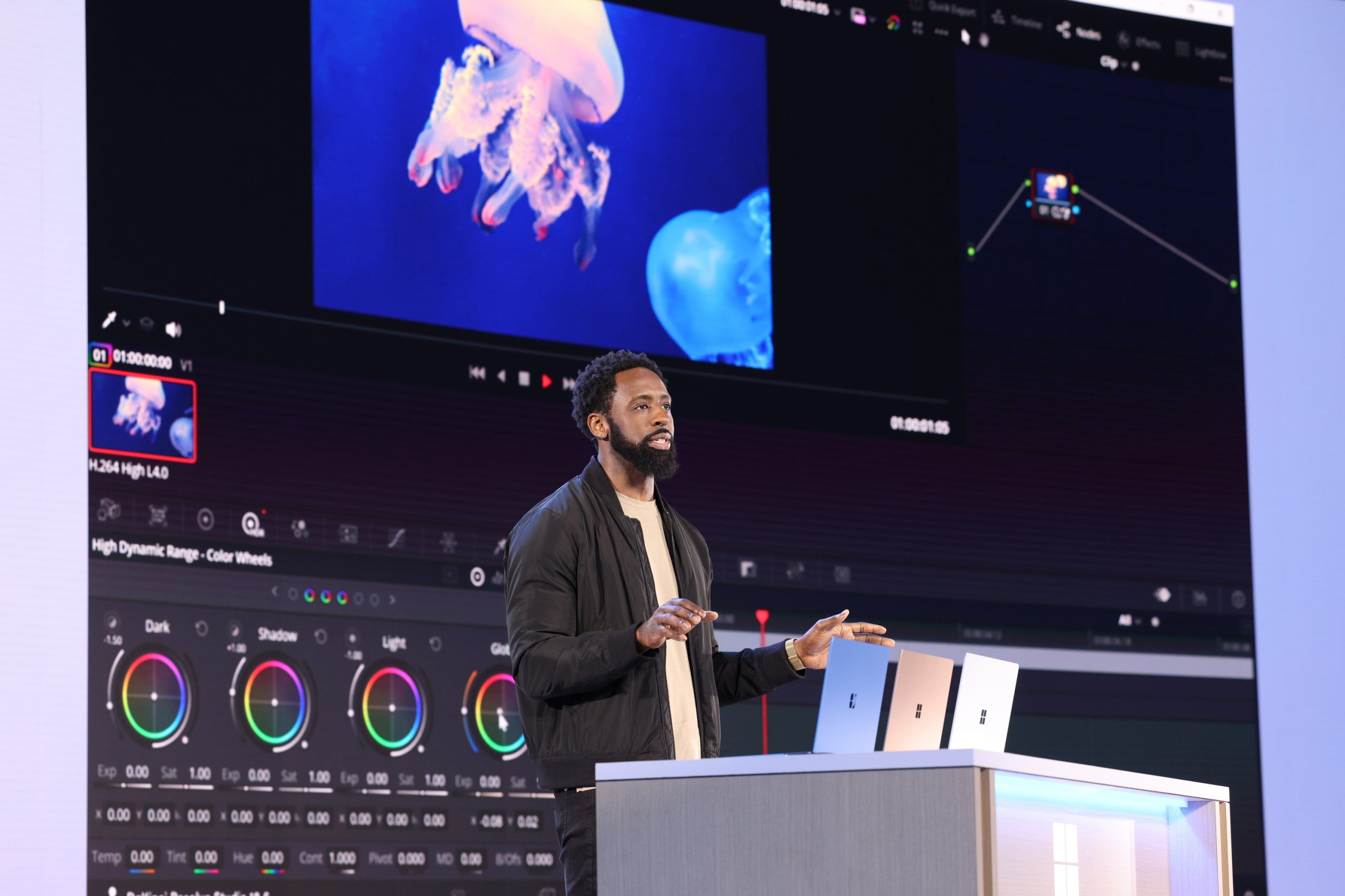 A man standing on stage in front of an audience with a product demonstration projected onto the screen behind him and three laptops on a table in front of him