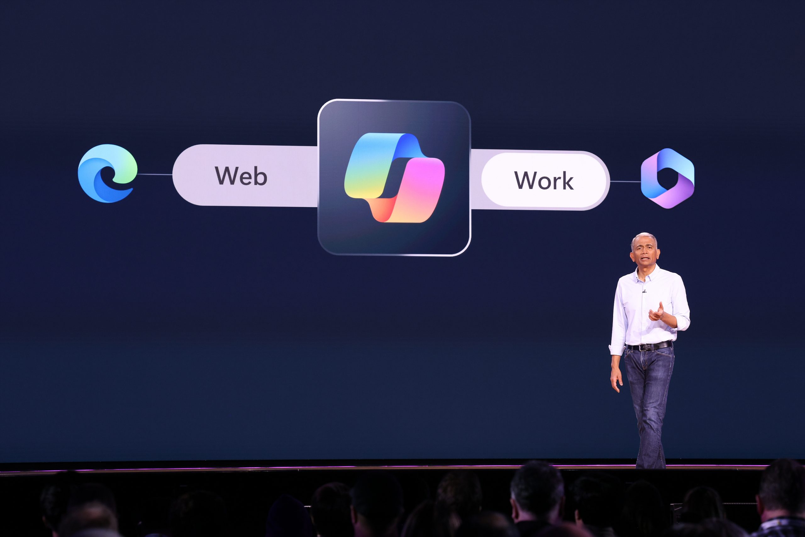 A man standing on stage with the Copilot logo and the words "Web" and "Work" on a screen behind him