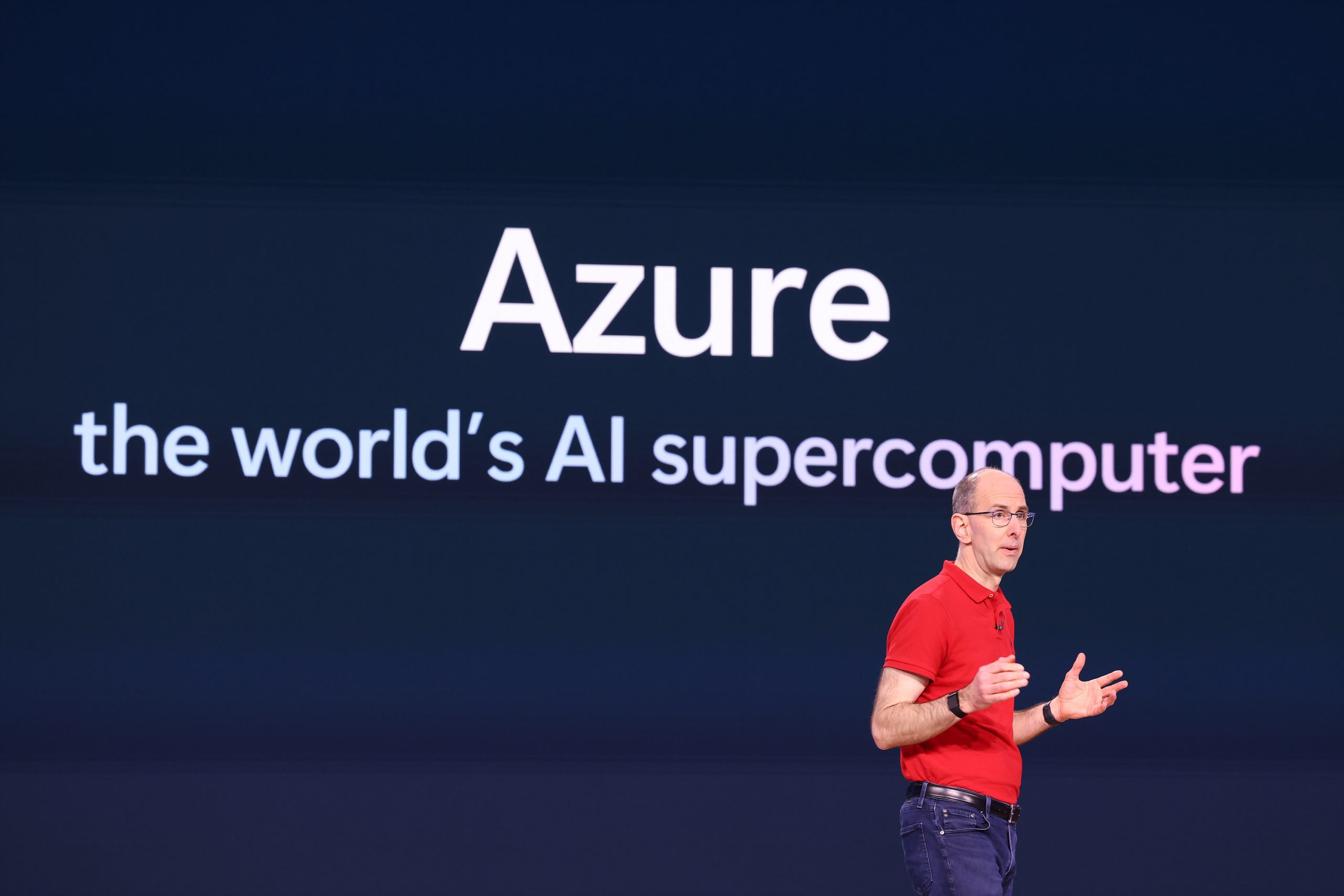 A man standing on stage speaking to an audience and gesturing with his hands, with text on the screen behind him that reads, "Azure, the world's AI supercomputer"