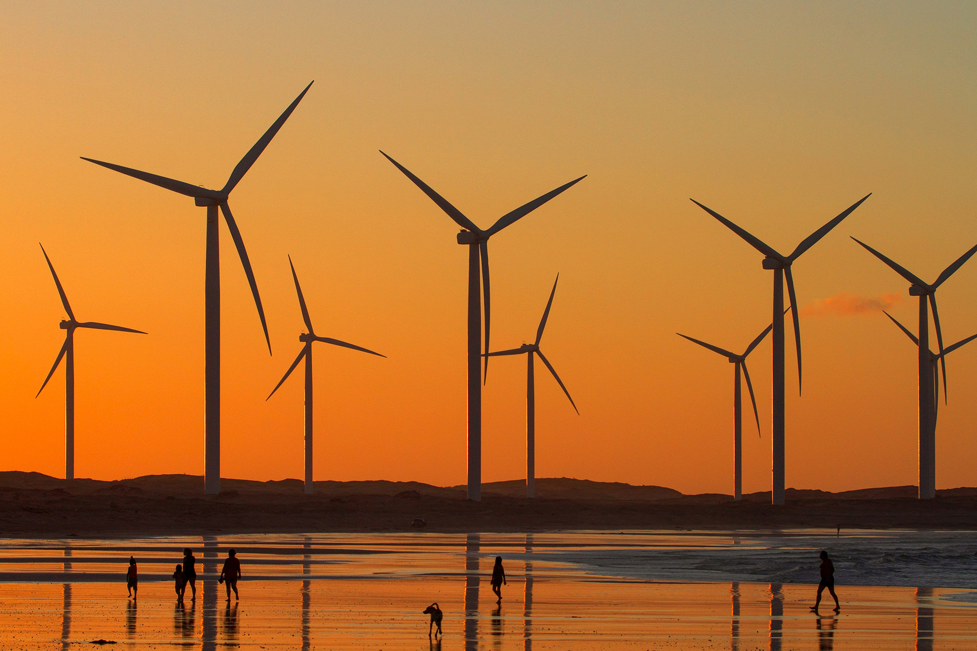 Silhouette of wind turbines on a beach at sunset, with people walking along the shoreline.