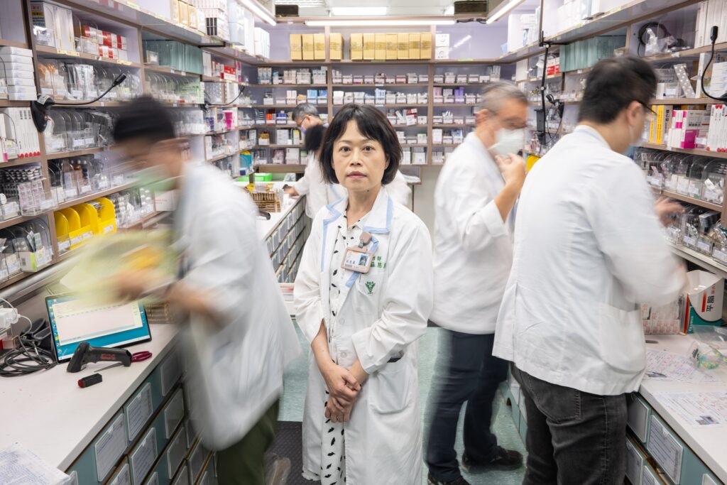 A healthcare professional standing in the middle of a busy hospital pharmacy, surrounded by other staff members working quickly in the background.