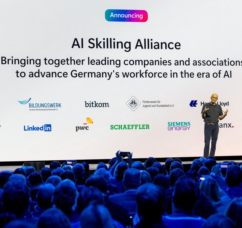 Satya Nadella steht auf der Bühne und hält eine Präsentation. Er trägt ein schwarzes Oberteil und dunkle Hosen. Hinter ihm ist ein großer Bildschirm, der die Worte zeigt: "AI Skilling Alliance. Bringing together leading companies and associations to advance Germany's workforce in the era of AI".