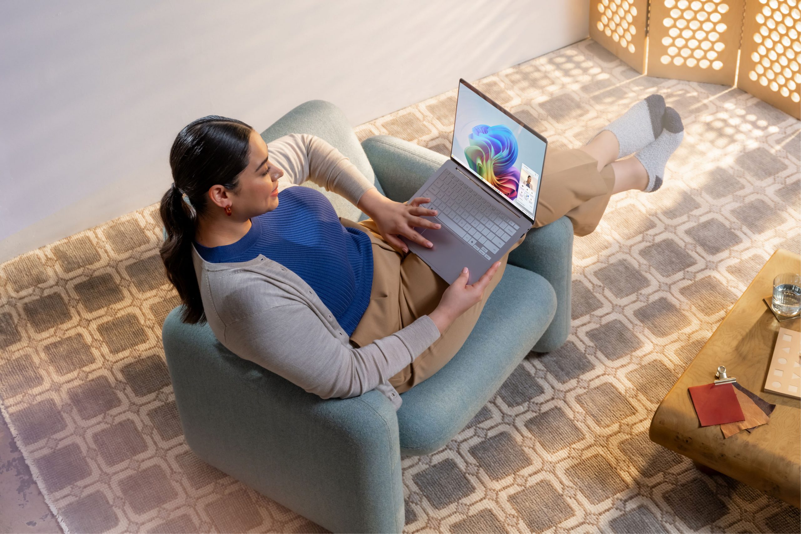 Photography showing an adult using a Copilot+ PC while casually sitting sideways in a soft, blue accent chair.