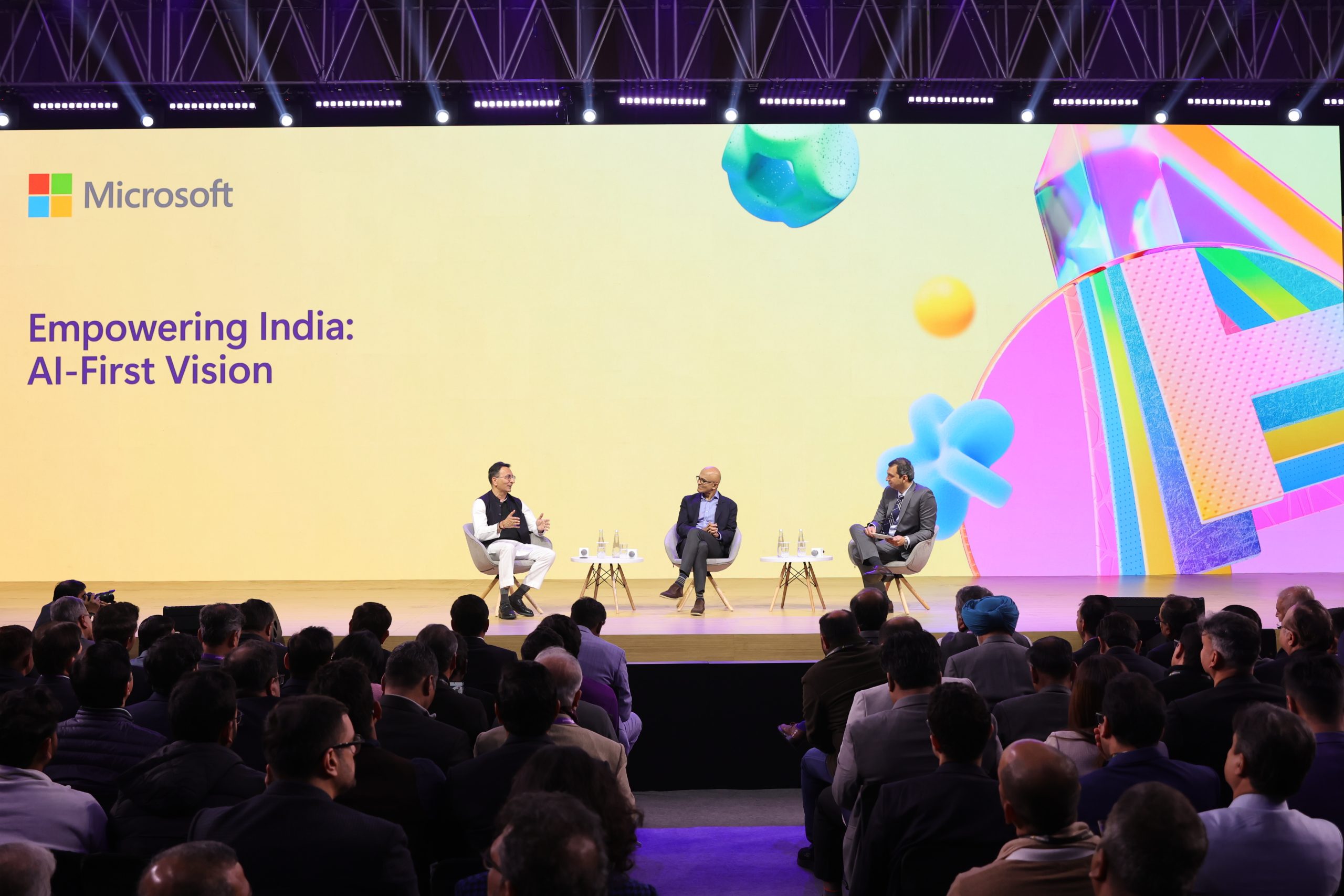 three men seated on stage engaged in conversation backdrop reads empowering india: AI first vision