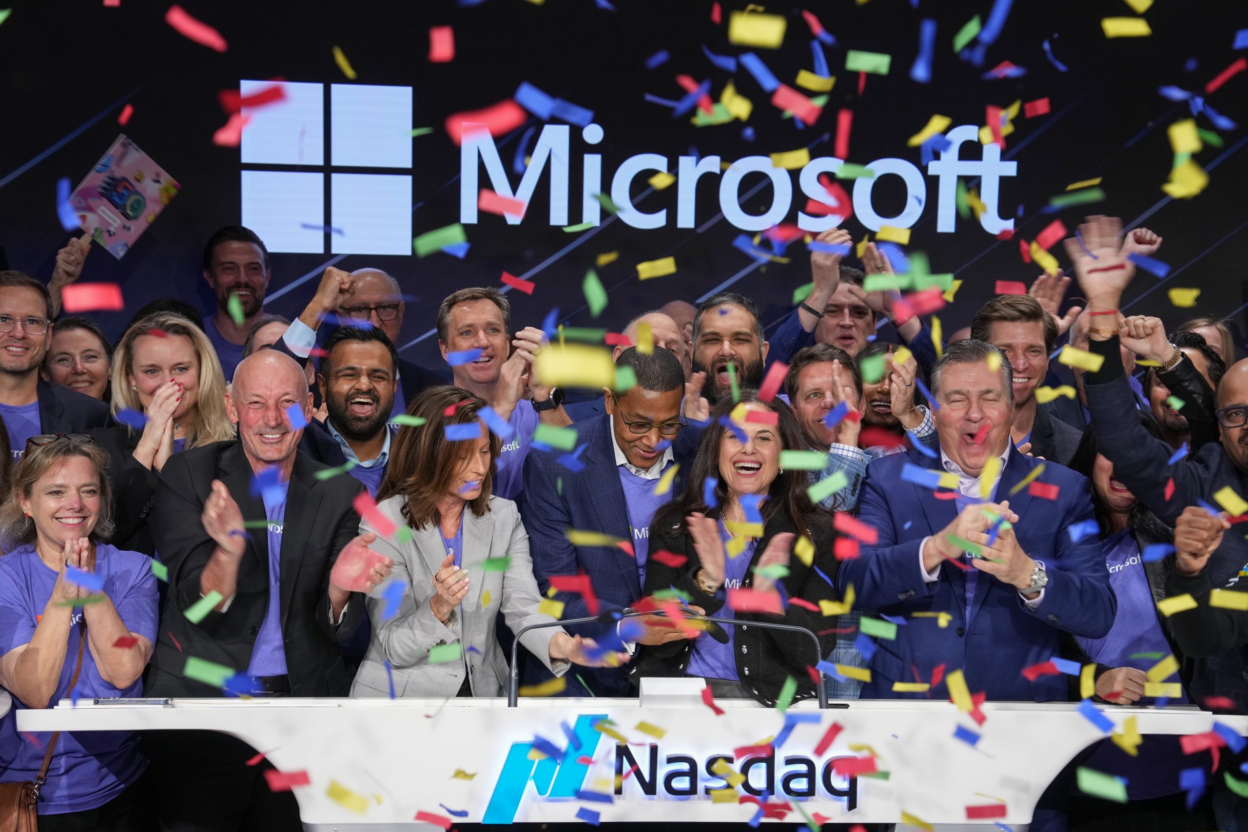 A group of people are celebrating on a stage at the Nasdaq MarketSite with colorful confetti falling around them. They are standing in front of a large screen displaying the Microsoft logo, and the Nasdaq logo is visible on the podium in front of them.