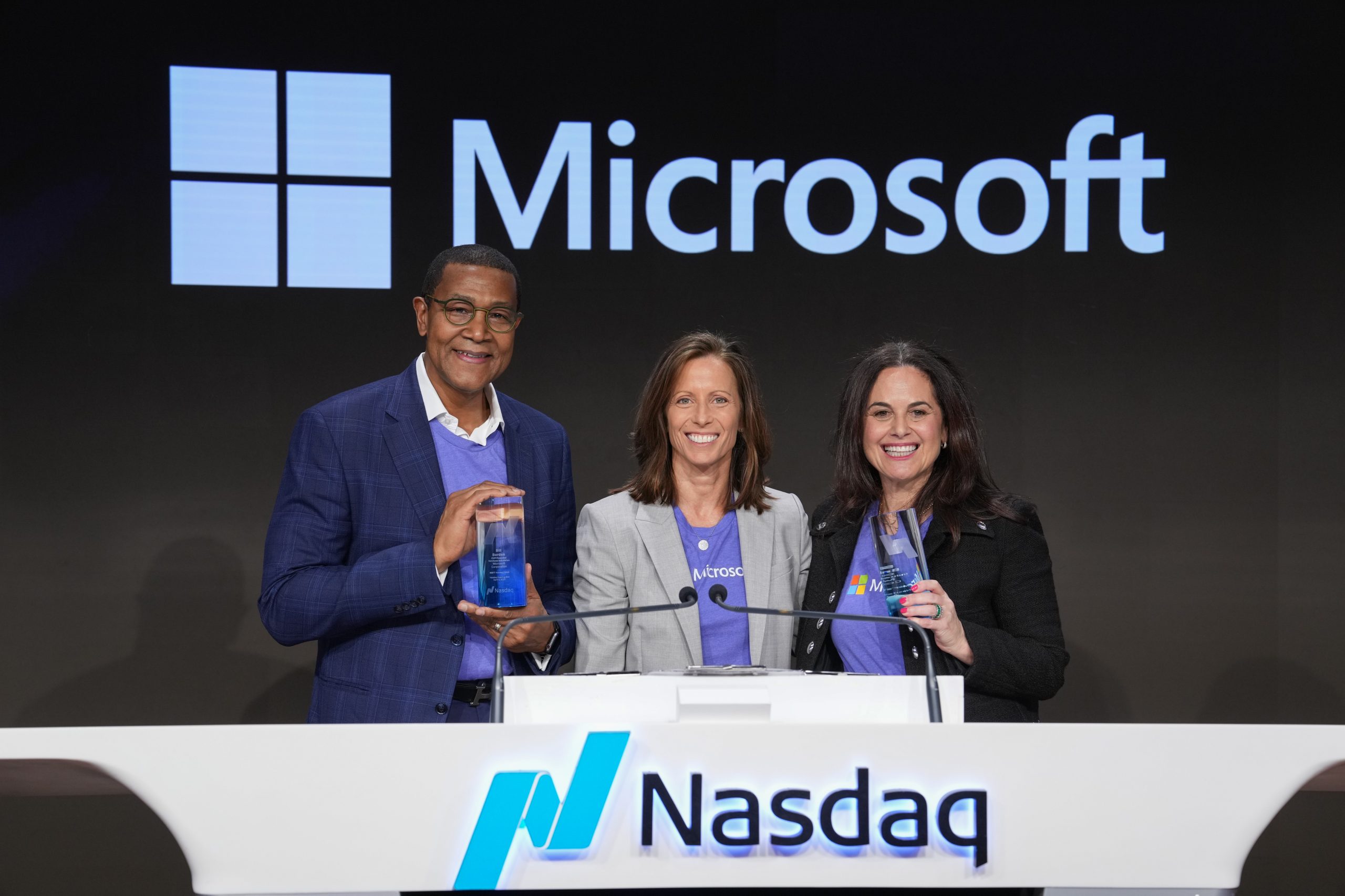 Three individuals stand behind a podium with the Nasdaq logo, two are holding glass awards. The background features a large Microsoft logo.