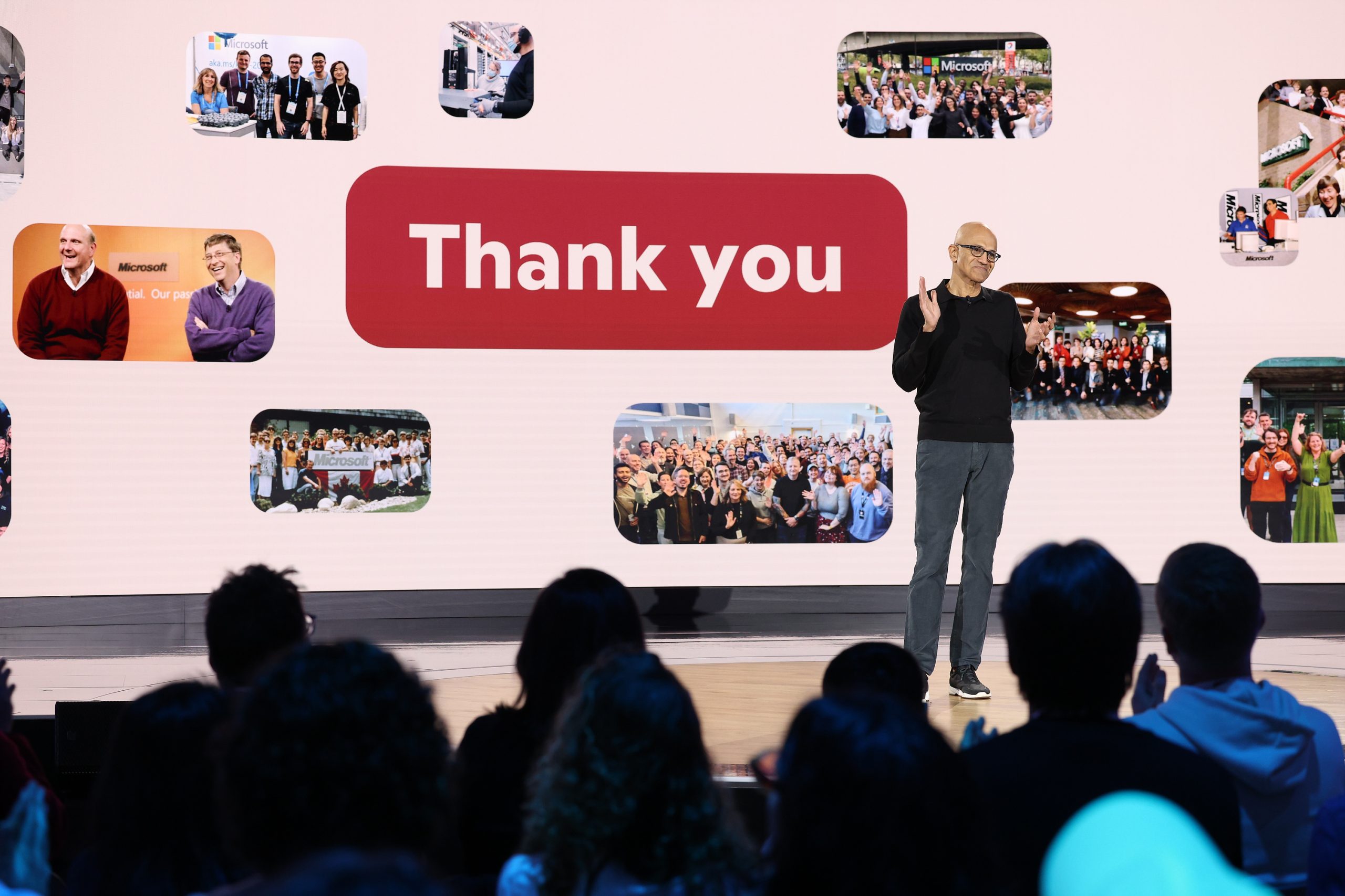 A man standing on a stage shares applause with an audience in front of a screen that reads, “Thank you.”