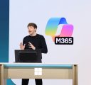 A man in a black sweater stands behind a computer onstage. Behind him is the Microsoft 365 logo on a large screen.