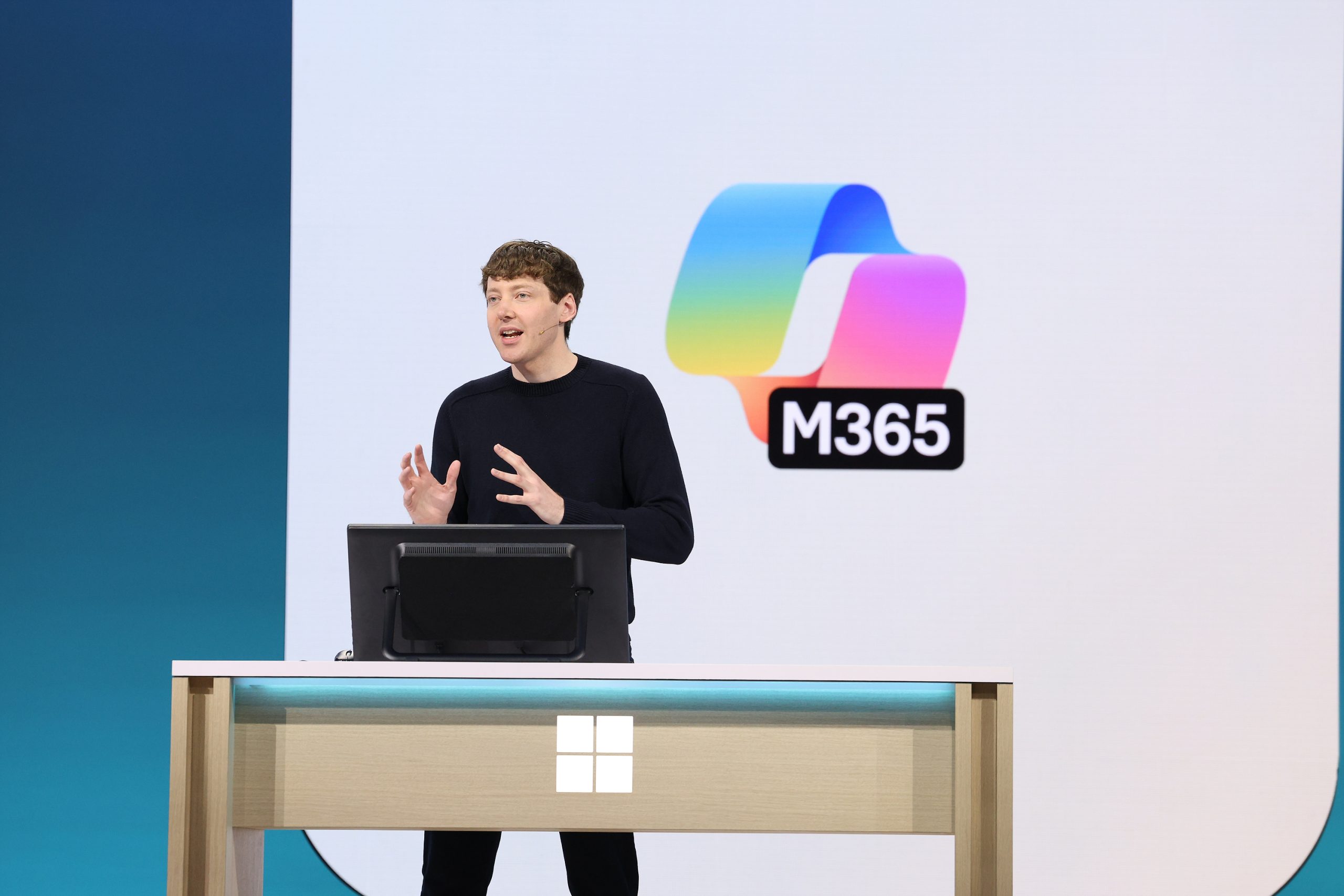 A man in a black sweater stands behind a computer onstage. Behind him is the Microsoft 365 logo on a large screen.