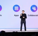 A man in a black sweater and jeans stands onstage in front of a large screen that reads, “agents, apps, collaboration.”