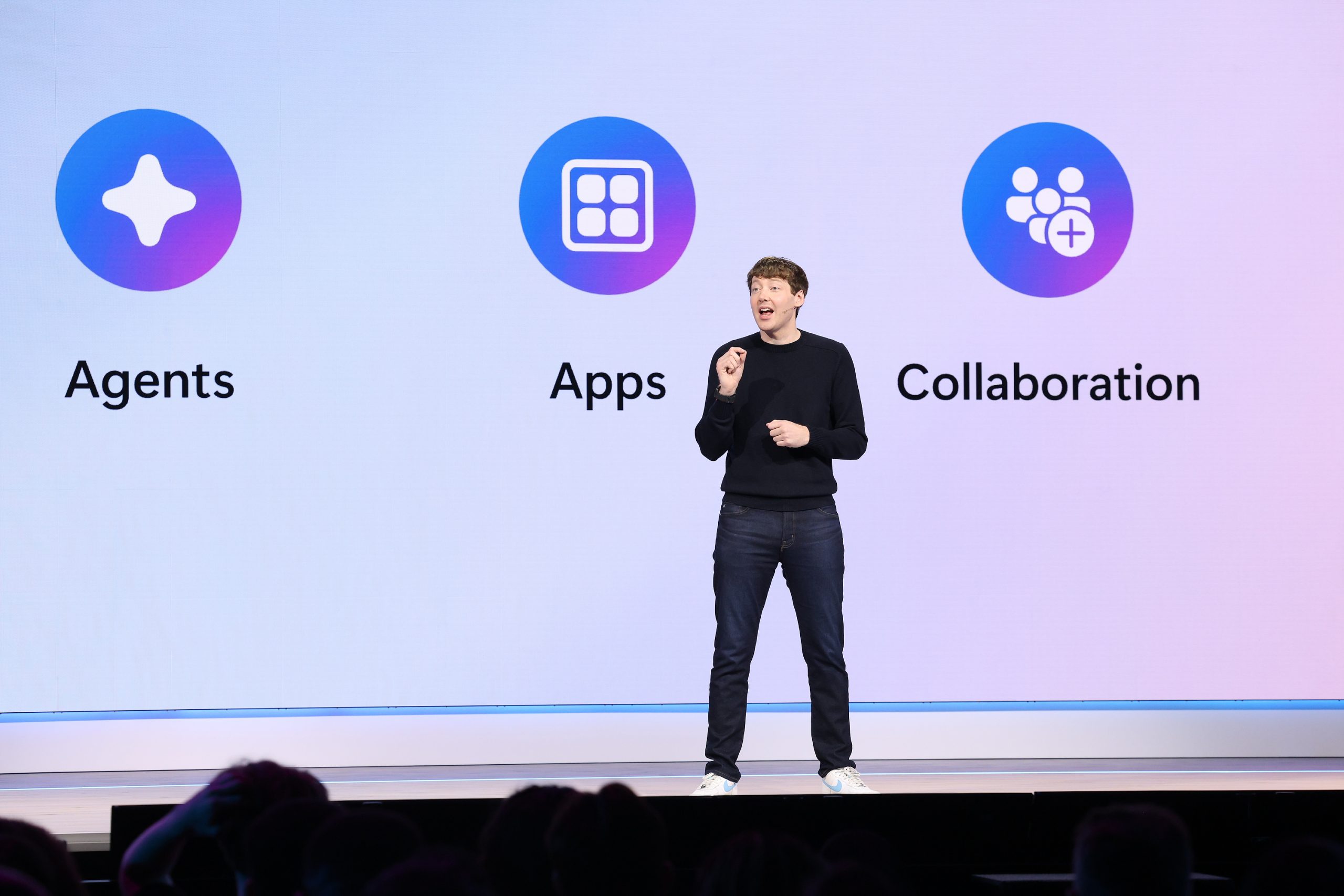 A man in a black sweater and jeans stands onstage in front of a large screen that reads, “agents, apps, collaboration.”