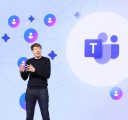 A man in a black sweater and jeans stands onstage in front of a large purple screen with the Microsoft Teams logo on it.
