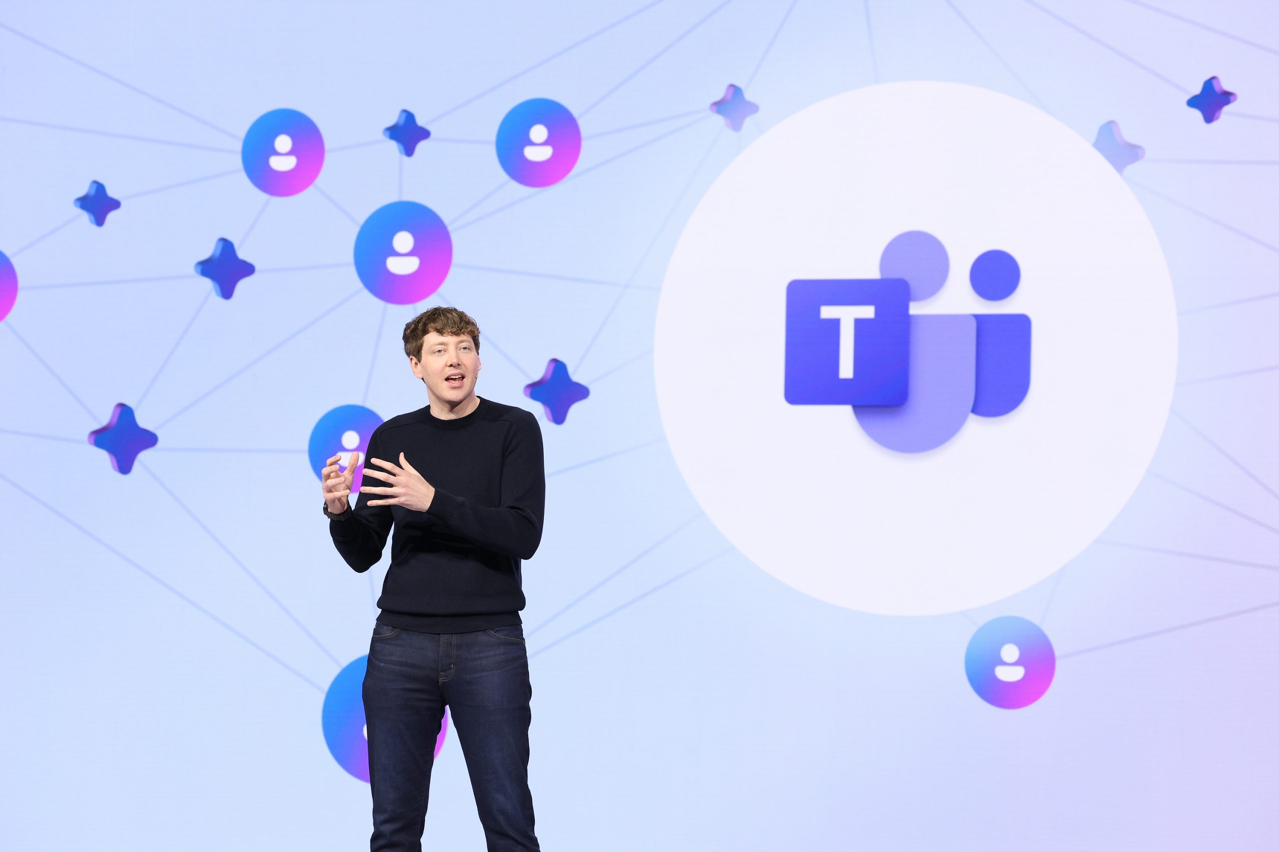A man in a black sweater and jeans stands onstage in front of a large purple screen with the Microsoft Teams logo on it.