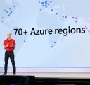 A man in glasses, a red polo and jeans stands onstage. He is in front of a large screen that reads, “70+ Azure regions.”