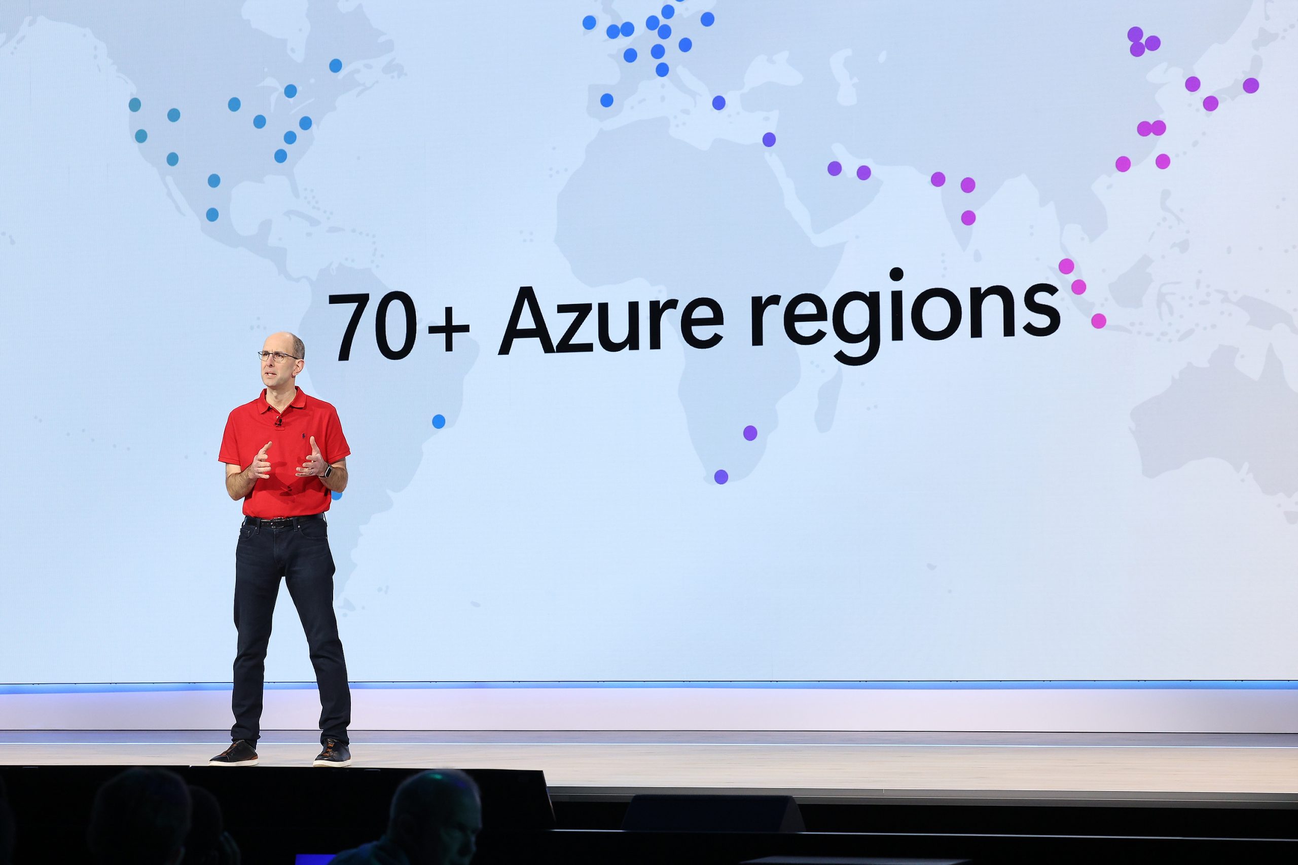 A man in glasses, a red polo and jeans stands onstage. He is in front of a large screen that reads, “70+ Azure regions.”