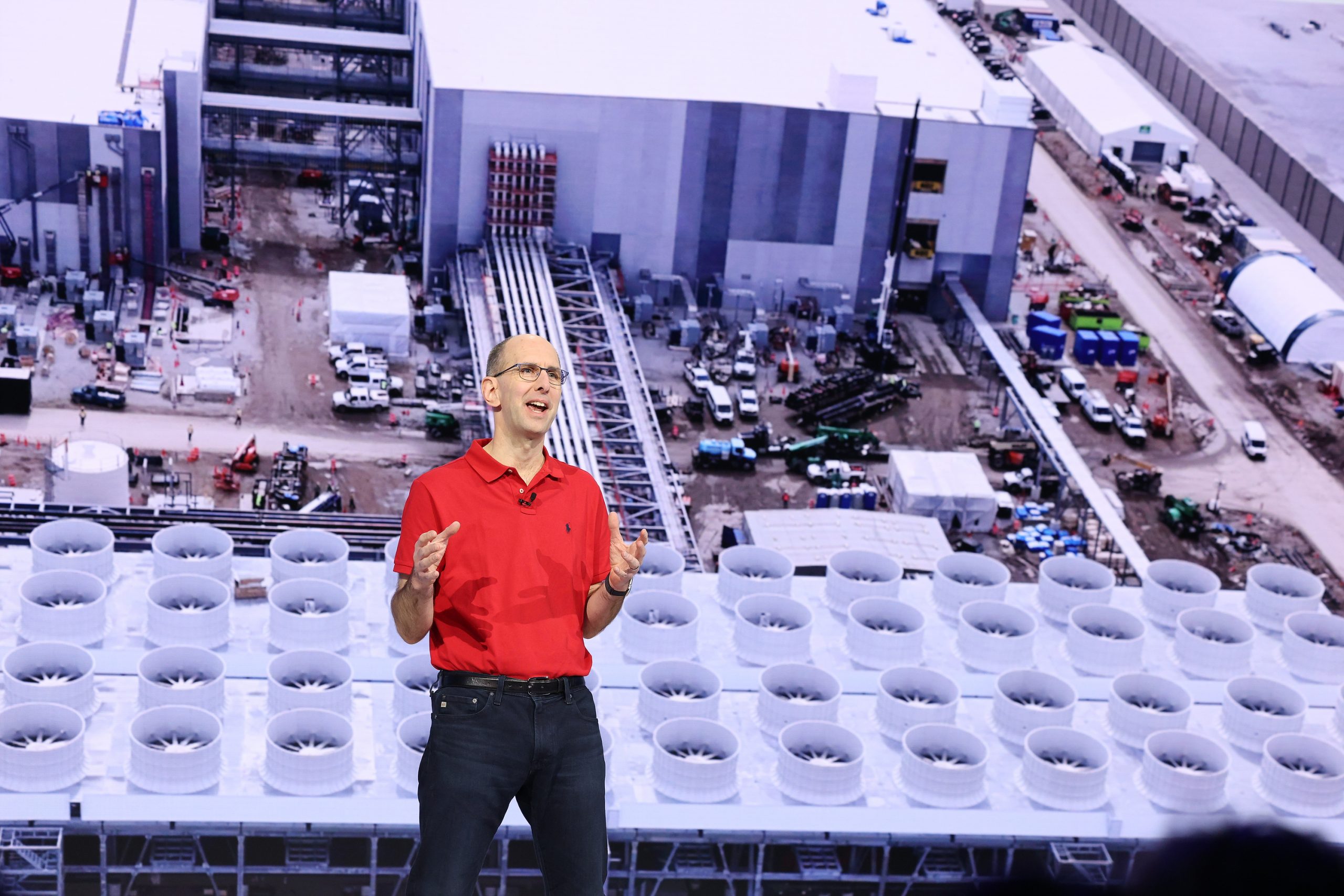 A man in a red polo, glasses and jeans, stands onstage. He is gesturing with his hands in front of a large screen with an image of a building.