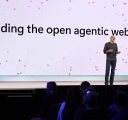 A man in jeans and a grey sweater speaks to an audience from a stage. He is in front of a large screen that says, “Building the open agentic web.”