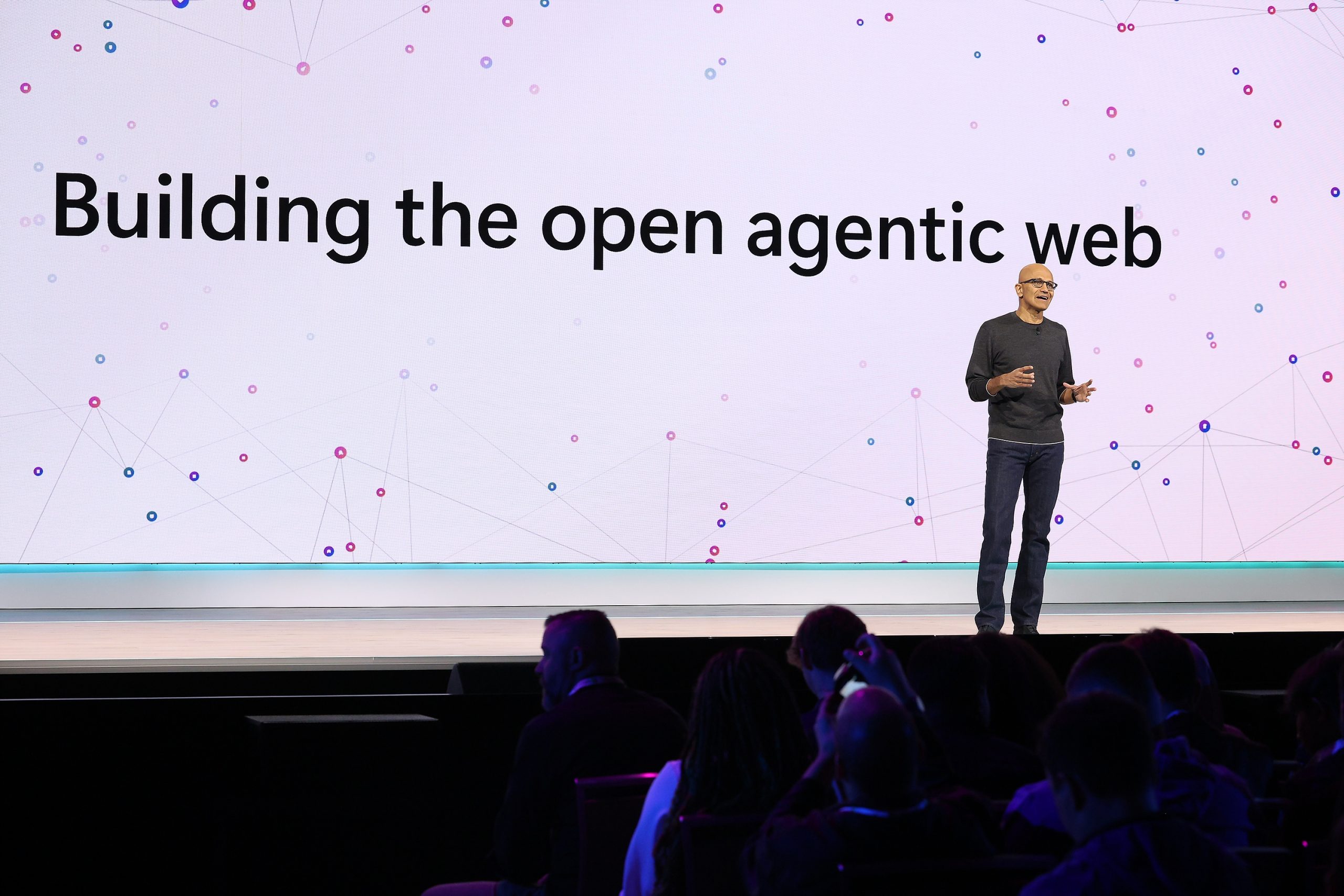 A man in jeans and a grey sweater speaks to an audience from a stage. He is in front of a large screen that says, “Building the open agentic web.”