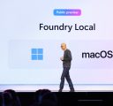 A man onstage in black glasses and a gray crewneck sweater walks in front of a large screen that reads “Foundry Local.”