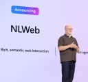 A man in glasses, a brown polo and jeans, stands onstage in front of a screen that says “Announcing NLWeb, rich semantic web interaction.”