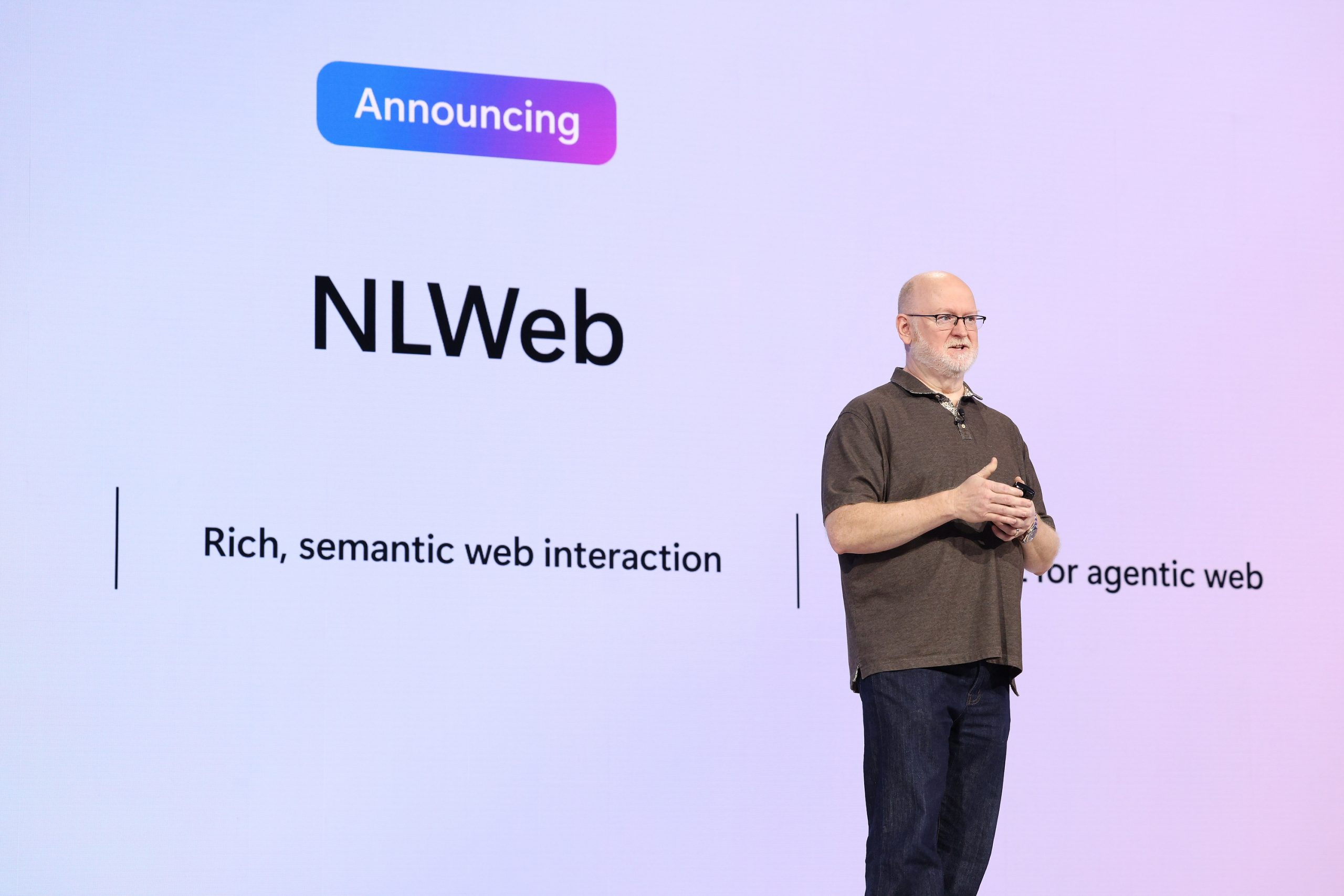 A man in glasses, a brown polo and jeans, stands onstage in front of a screen that says “Announcing NLWeb, rich semantic web interaction.”