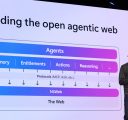 A man in a dark grey sweater stands onstage in front of a large monitor with a flow chart. The onscreen title says, “Building the open agentic web.”
