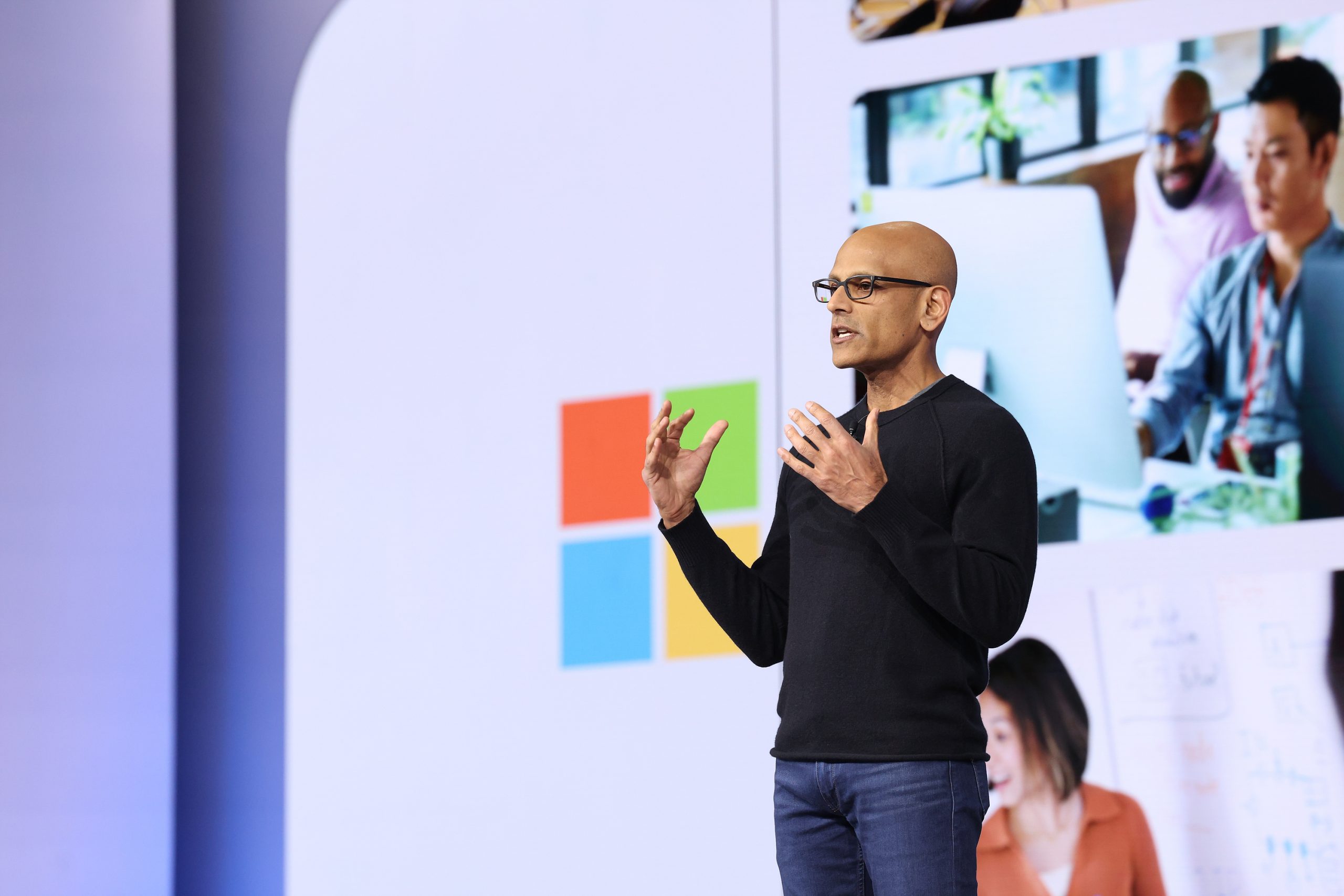 A man in black frame glasses, a black sweater and jeans stands onstage, speaking to an unseen audience.