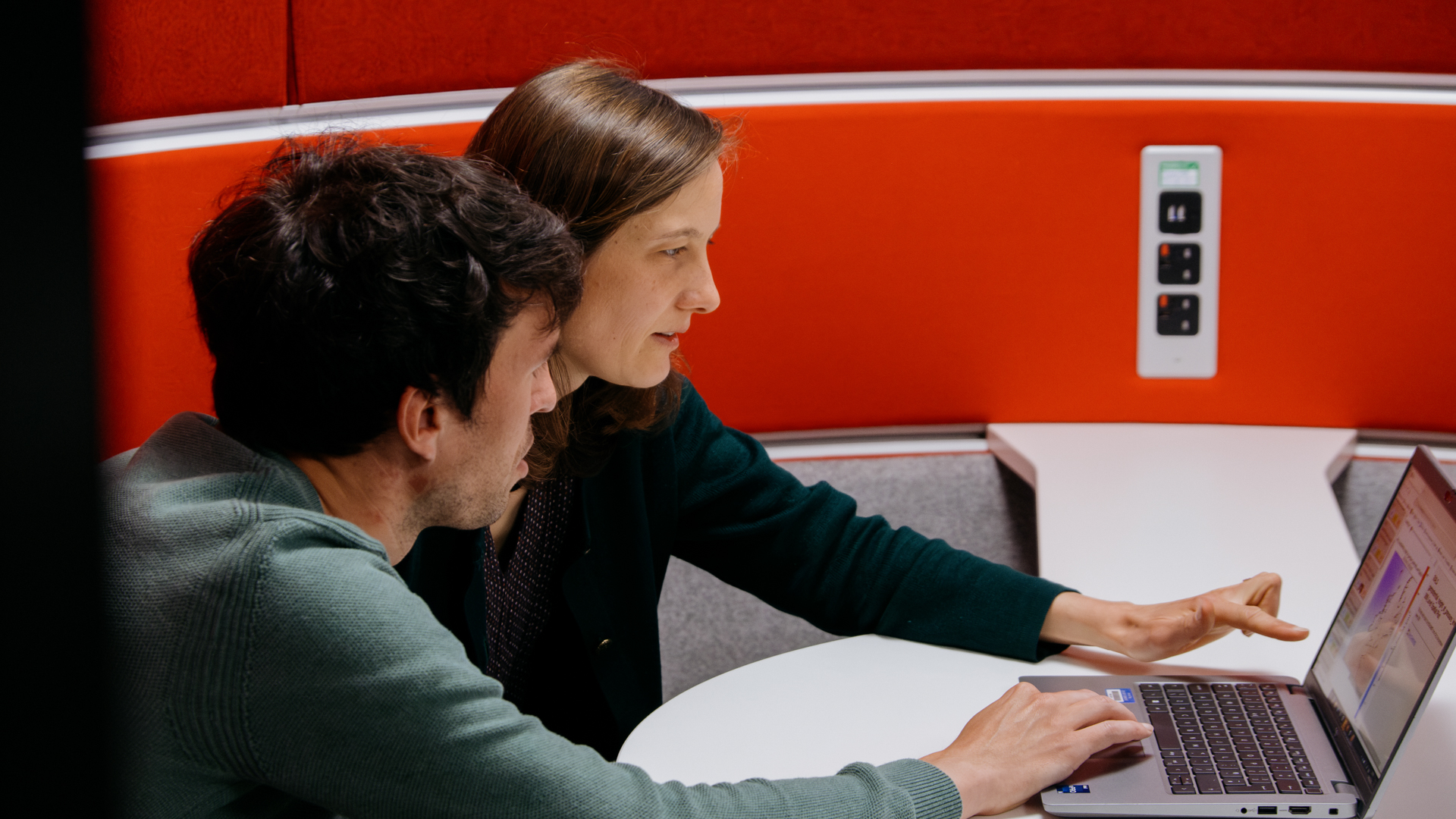 A woman with shoulder length light brown hair and a man with dark brown hair sitting at a table together looking at a laptop.