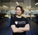 A man with an asymmetrical haircut, wearing a black T-shirt that says Stanford Medicine, stands with arms crossed in a glass office space.