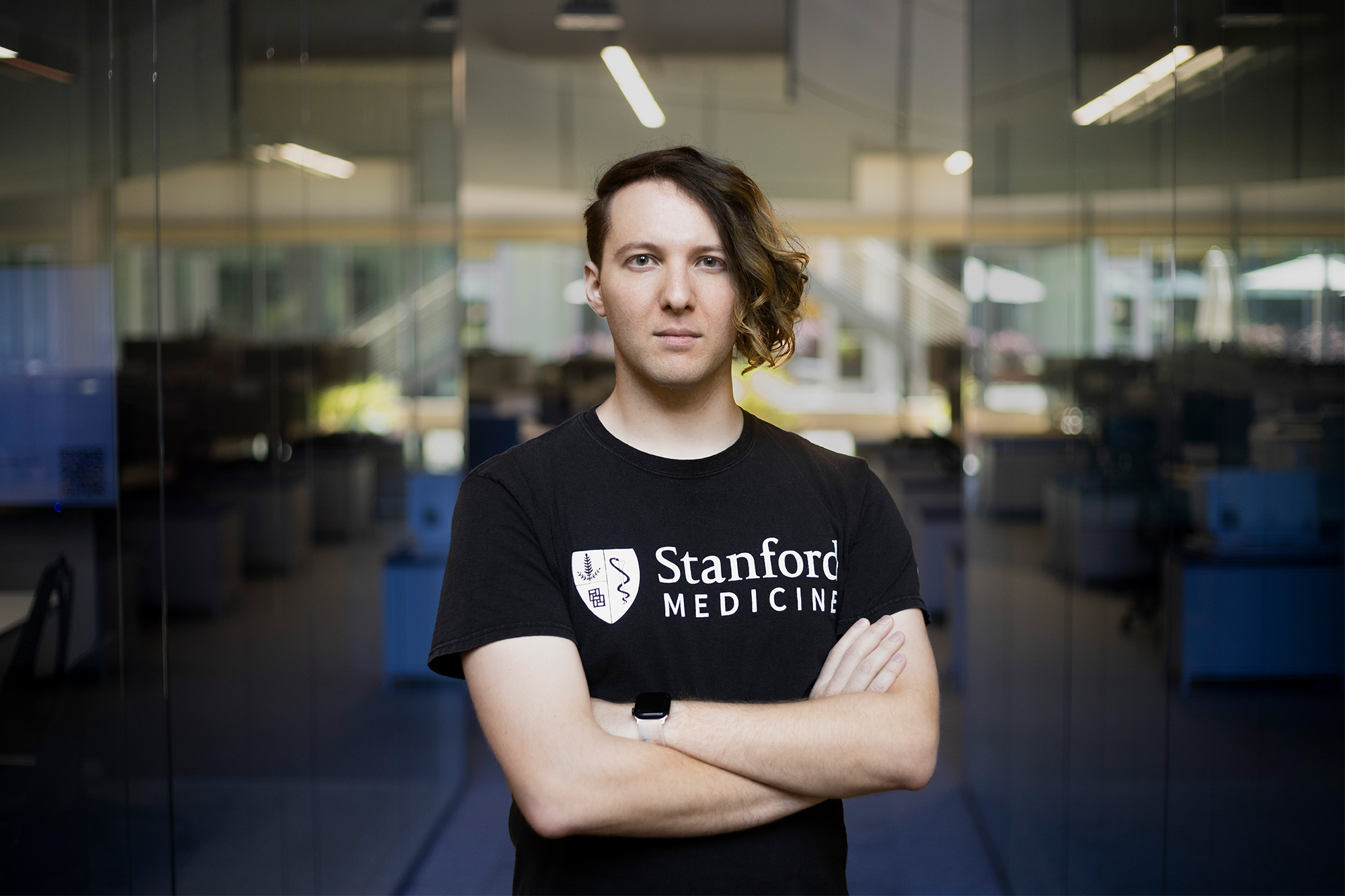 A man with an asymmetrical haircut, wearing a black T-shirt that says Stanford Medicine, stands with arms crossed in a glass office space.