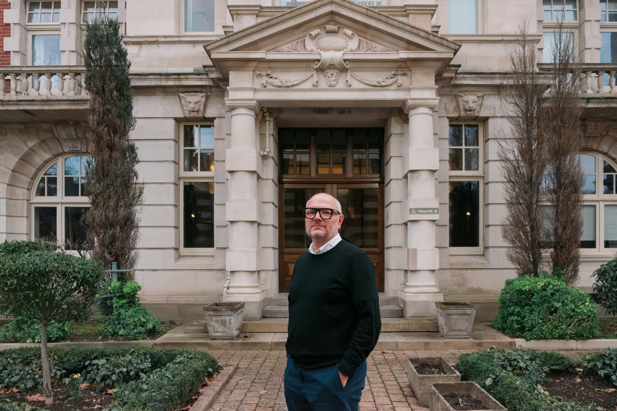 Man in black framed glasses in front of an ornate brick building.