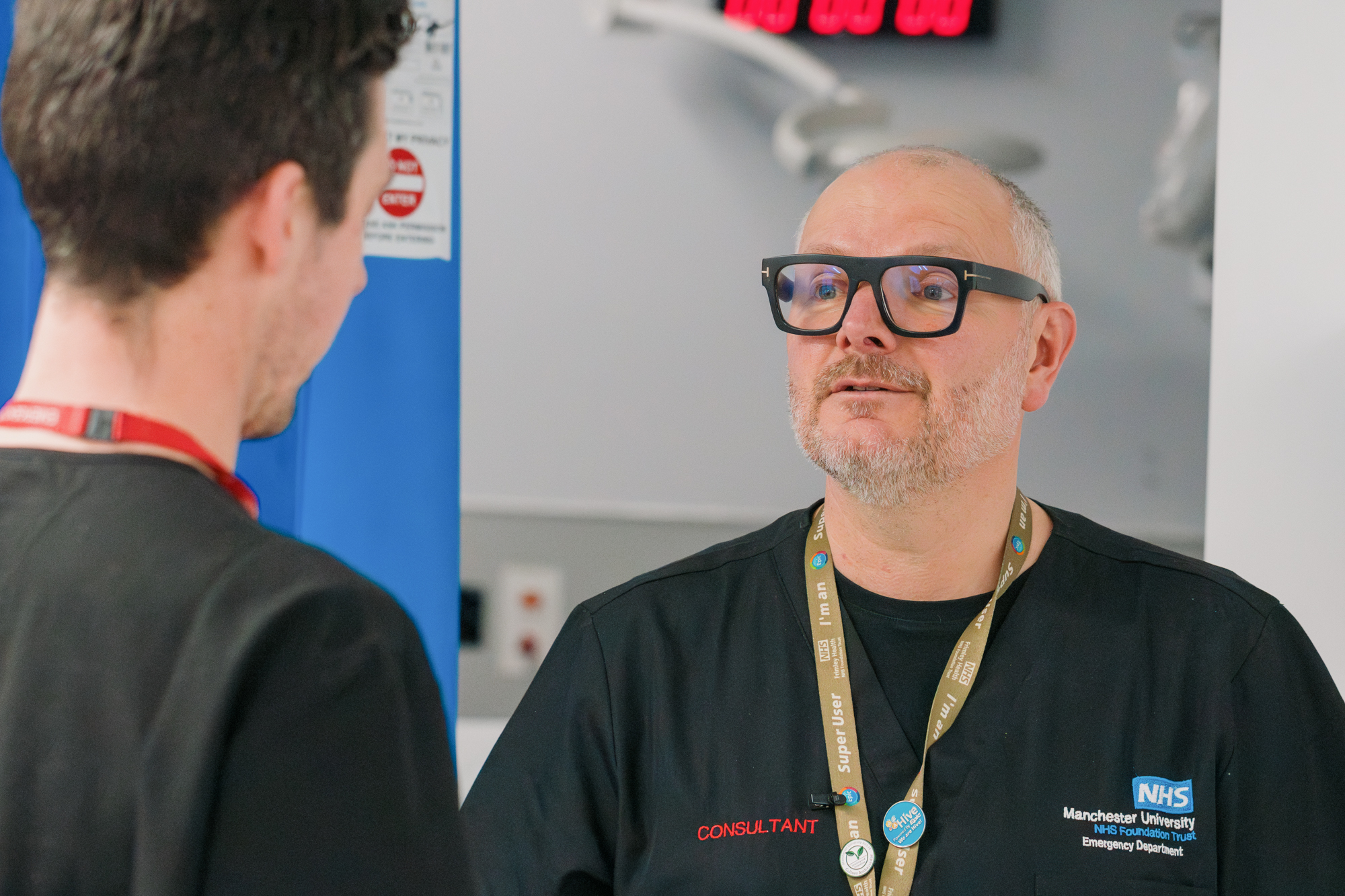 A man in black framed glasses and black hospital scrubs speaks with another man who is seen from behind, also wearing hospital scrubs.