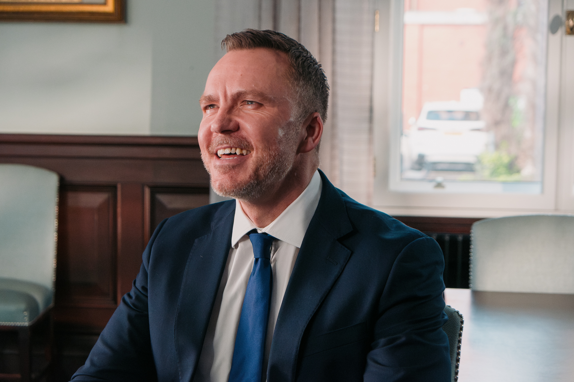 Portrait of a smiling man in a chair wearing a blue suit with a white shirt and lighter blue tie.