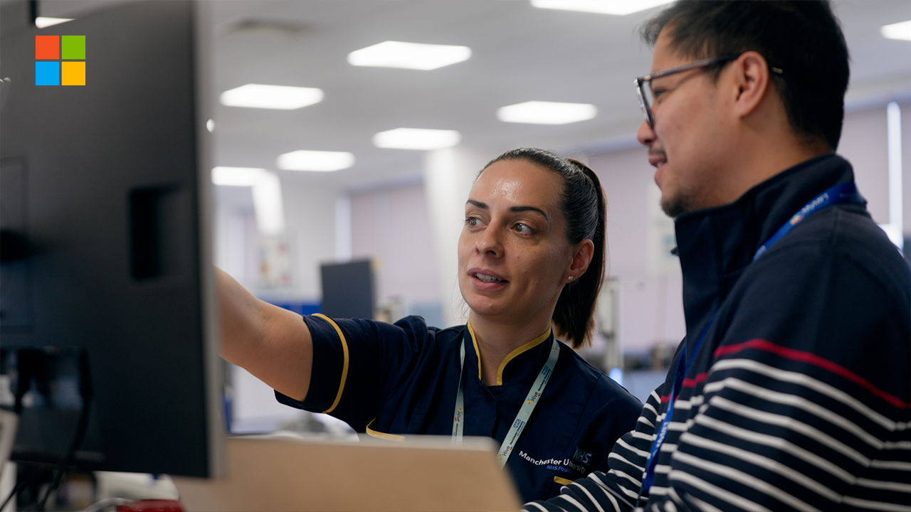 Two NHS staff members standing in a clinical office environment, reviewing information together on a large computer screen, with NHS uniforms and lanyards visible.