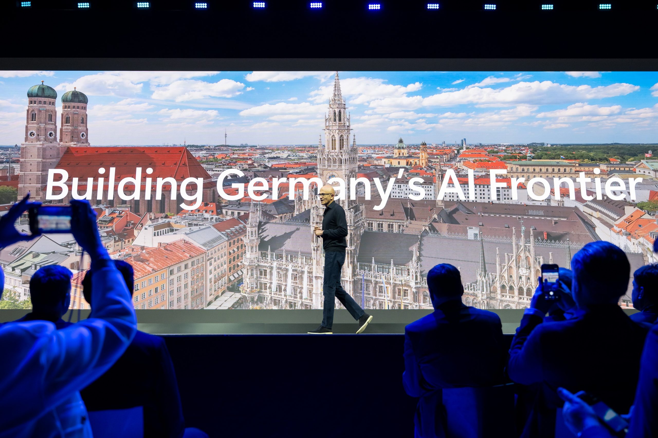 Redner auf der Bühne vor Münchner Skyline und dem Schriftzug „Building Germany’s AI Frontier“ mit Publikum im Vordergrund.